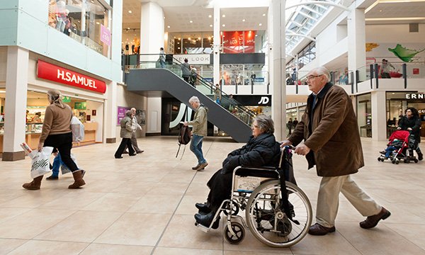 woman in wheelchair being pushed through shopping centre by man