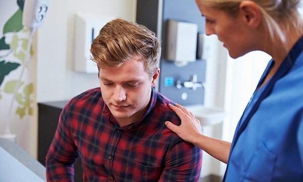 Picture shows a young man in a hospital room being reassured by a nurse, who has a hand on his shoulder. This article stresses the importance of investing in mental health nursing leadership on acute medical and surgical wards.