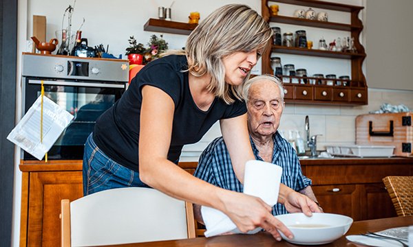 woman helps frail man to take a meal at a kitchen table