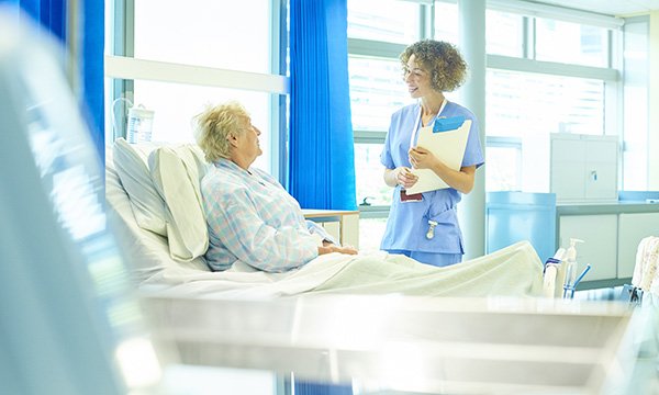 Picture shows a female medic chatting to an older woman who is sitting up in a hospital bed. The article says the nursing profession needs to debate whether intentional rounding is the best way to deliver care to patients.