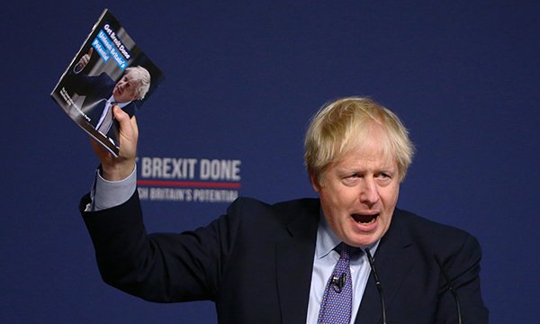 Prime minister Boris Johnson speaks at a podium, holding aloft the Conservatives' 2019 election manifesto