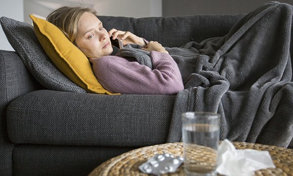 woman lies on sofa under blanket, glancing at tablets and a glass of water of the coffee table next to her