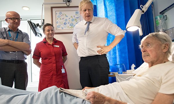 Prime minister Boris Johnson speaking to a patient on a ward, accompanied by healthcare staff