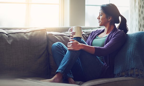 woman sits on sofa, mug in hand, looking out of a window