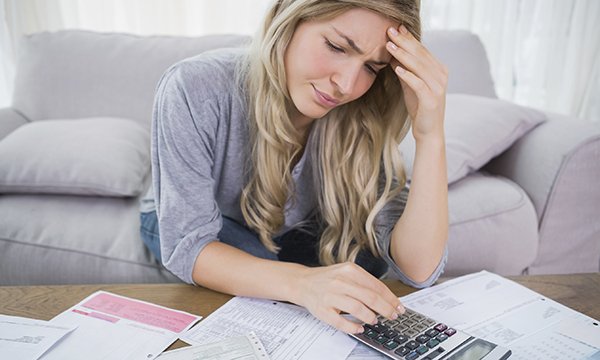 worried-looking woman sits on sofa at home, working with a calculator as she pores over financial statements
