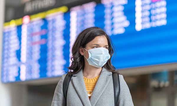 Picture shows a woman wearing a face mask, with an airport departures board in the background