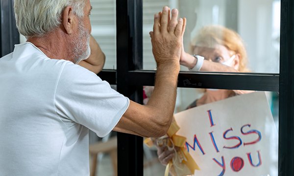 Picture shows an older couple separated by a window and holding their hands against the glass to greet each other