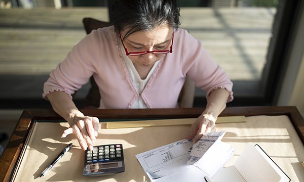 Picture shows a woman sitting at a desk leafing through bills while using a calculator