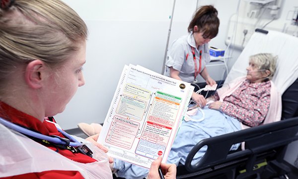 Picture shows a nurse at the bedside of an elderly patient while another nurse looks at a sepsis checklist