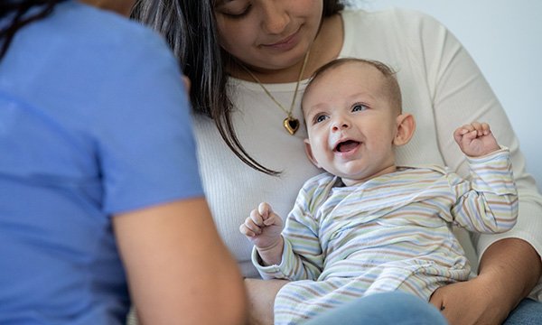 Photo shows nurse looking at a smiling baby in its mother's arms: backlogs of care and the unmet needs of families with babies and children are on the rise but overlooked