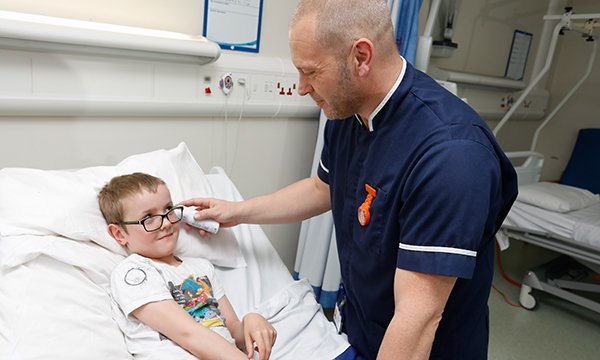 A male nurse chats and jokes with a boy who is in bed in a paediatric assessment unit