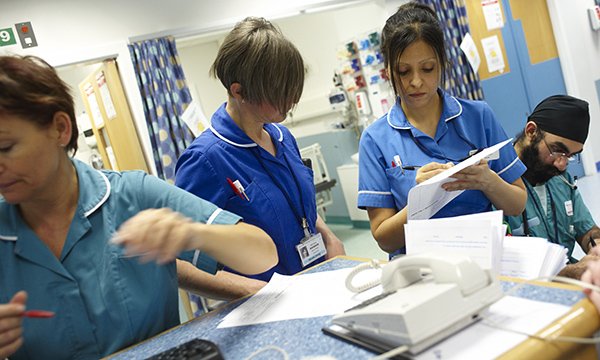 Four nurses carrying out tasks around a busy work station, despite recruiting 800 extra overseas nurses it has failed to reverse the decline in numbers of the NHS workforce