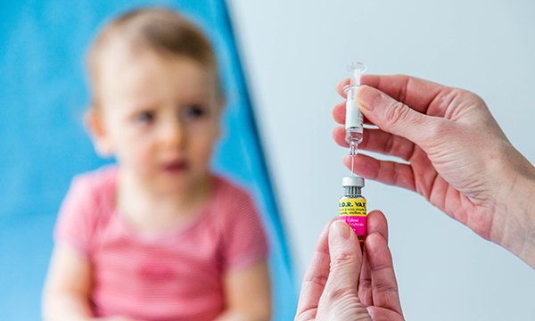Hands drawing a vial of a vaccine into a needle in the foreground, with a blurred image of a child in the background