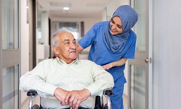 An older man in a wheelchair chats to a smiling nurse behind him