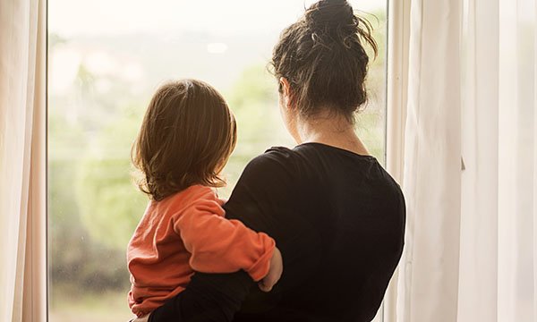 A woman holds a child in her arm as they both look out of the window