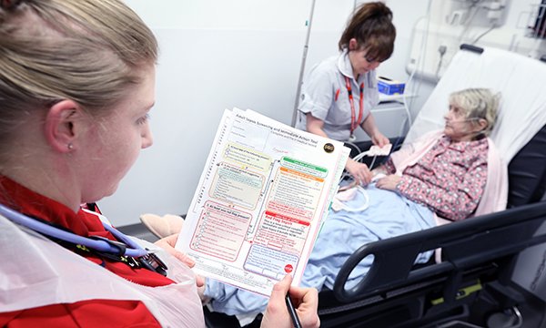 A medic standing at the bedside of an older patient checks a sepsis screening tool