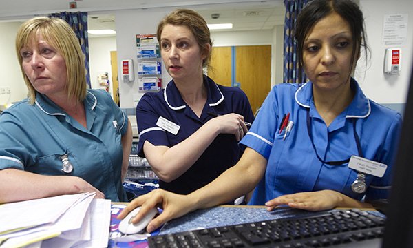 Three nurses stand cramped together at nurses' station, one is using a computer – as RCN points to gap between NHS waiting list and growth in nurse numbers