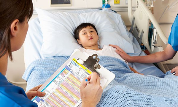 A nurse standing at the hospital bedside of a young child is watched by the child as she holds a file on which she is filling out a paediatric early warning system chart