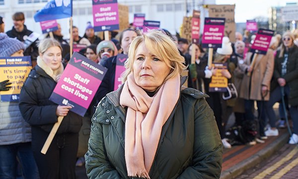 RCN general secretary looks serious during visit to nurses' picket line outside Leeds General Infirmary in December 2022. Behind her, striking nurses hold signs with messages about staffing and pay. Everyone is dressed for winter weather