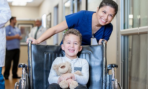 A young boy smiles and hugs a teddy bear while being pushed in a wheelchair along a hospital corridor by a smiling nurse