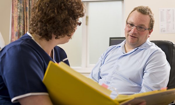 A nurse holds open a folder to show something to a patient sitting opposite
