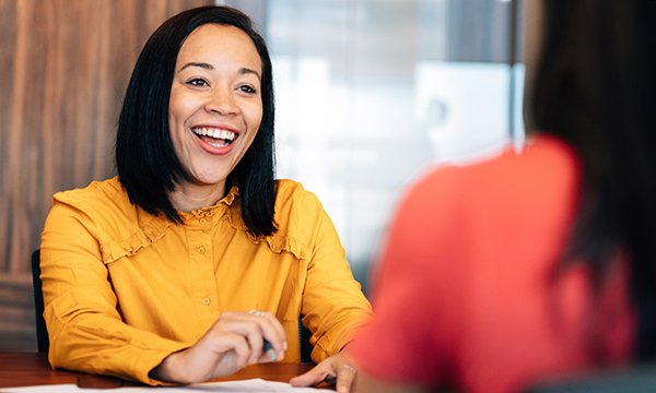 A woman in a smart yellow blouse smiles widely at the person sitting opposite her at a desk, as in a nursing job interview
