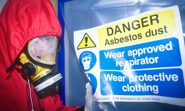 A figure wearing a protective suit and mask covered with dust standing next to a sign warning of the danger of asbestos dust