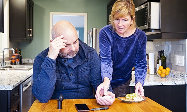 A worried-looking man at a kitchen table with a woman standing next him.
