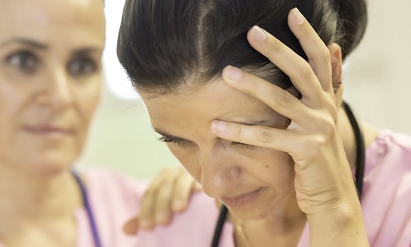 A nurse clutches a hand to her forehead while a concerned colleague places a hand on her shoulder