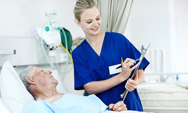 A nurse with a clipboard talks to an elderly patient in a hospital bed