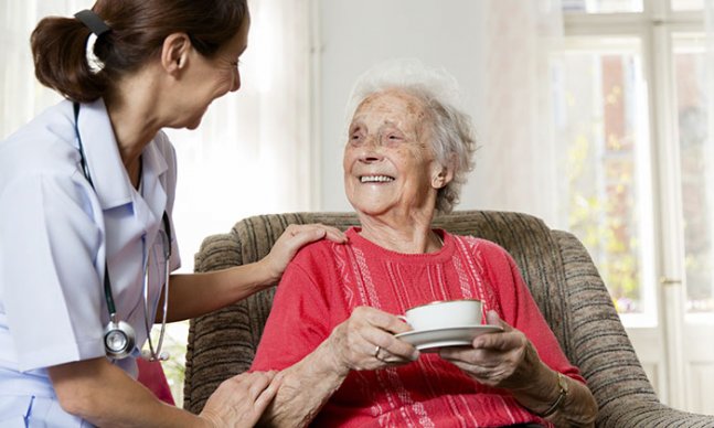 nurse sits beside a woman who is sitting in an armchair, holding a cup and saucer.They smile at each other