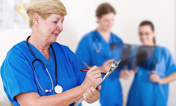Older nurse makes notes on a clipboard, with younger nurses standing behind her, viewing a lung X-ray