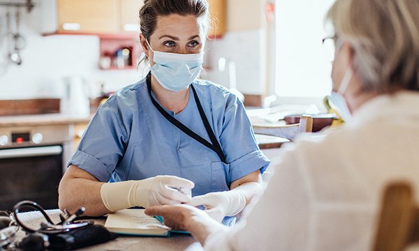 Picture shows nurse wearing a mask and gloves in a home visit with a female patient