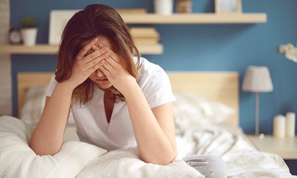 Woman sits slumped in bed, head in hands. Fatigue is among the symptoms of long-COVID