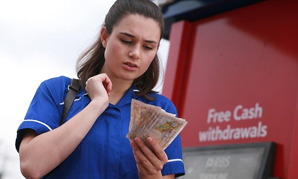 in nurse's uniform frowning as she counts money at a cash machine