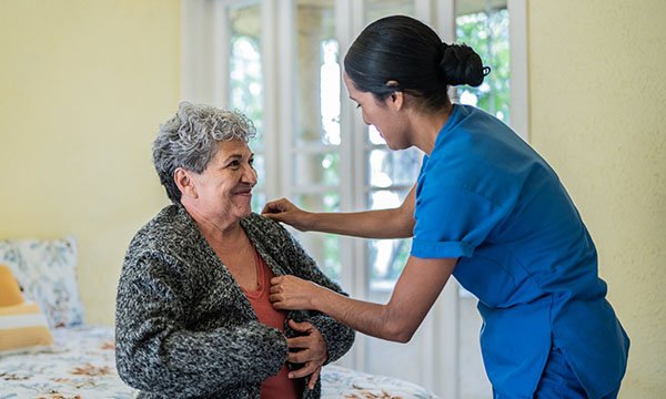 A nurse helps an older woman put her cardigan on in a care home setting