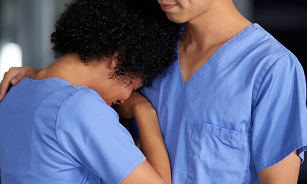 A distressed nurse leans against the shoulder of a colleague and is comforted