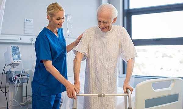 A nurse supports an older male patient by holding his shoulder as he grips a walking frame while moving away from his hospital bed