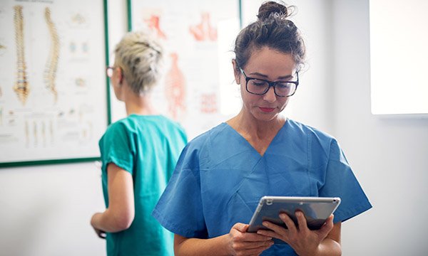 A clinician studies a handheld tablet while a colleague behind her looks at a medical wall chart