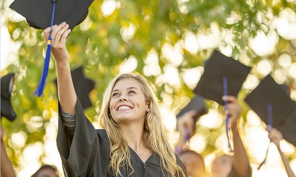 A smiling woman in a graduates gown holds her cap up in the sunlight