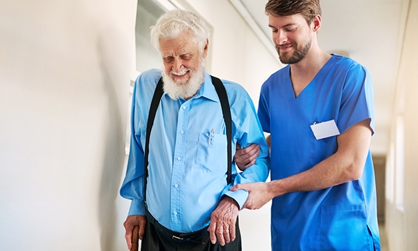 A nurse helps an elderly man walking down a corridor