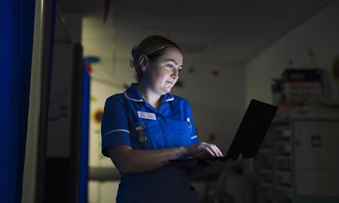 A nurse standing at a computer in a darkened room during a night shift, her face highlighted by the glow of the screen