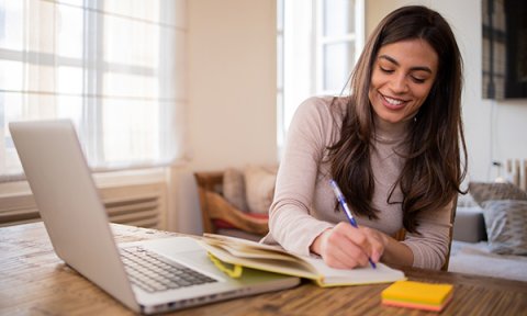 Woman smiles as she makes notes in front of her laptop while working at home – doctoral research is a time commitment and work-life balance challenge