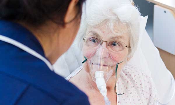 Picture shows an older woman in bed, wearing an oxygen mask, talking to a medic. Lorraine Creech describes the challenges facing the charity supporting people with mesothelioma