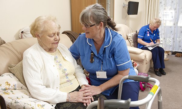 Seated woman in a nursing home sitting room is offered support by member of nursing staff