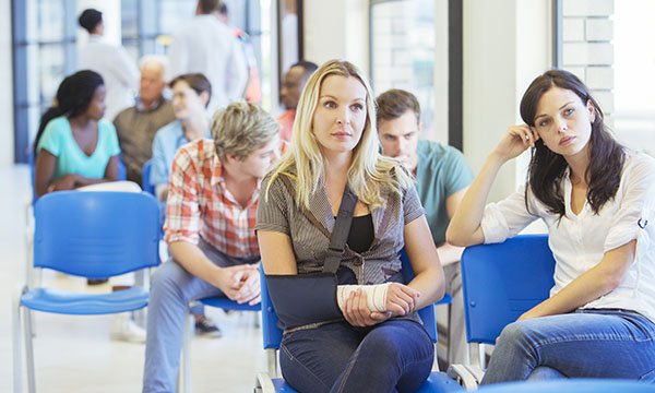People waiting in an emergency department waiting room, the front two women (one with her arm in a sling) look bored and fed up after a long wait