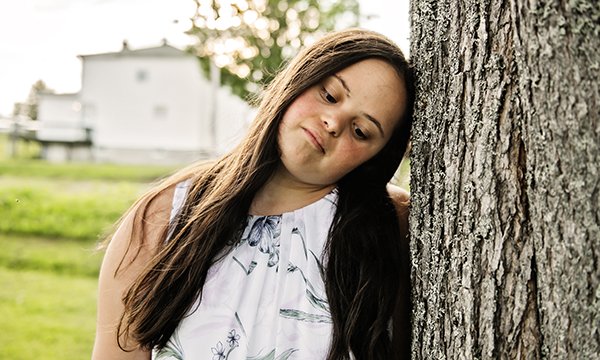 A  woman with Down’s syndrome leans her head against a tree looking downcast and vulnerable