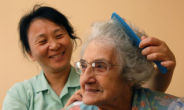 A smiling care worker combs the hair of an older woman who is looking content as she looks into the distance
