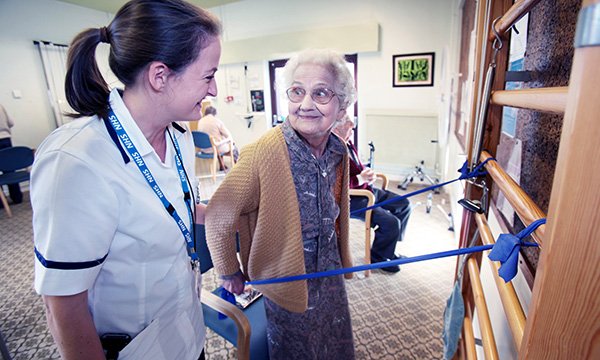 An older woman turns to a nurse and smiles during a falls assessment that involves holding lengths of tape fastened to wall bars