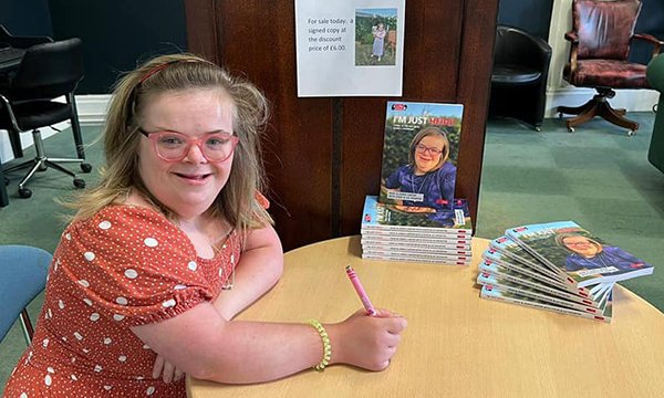A smiling Heidi Crowter sitting at a table with pen in hand ready to sign copies of her book I’m Just Heidi